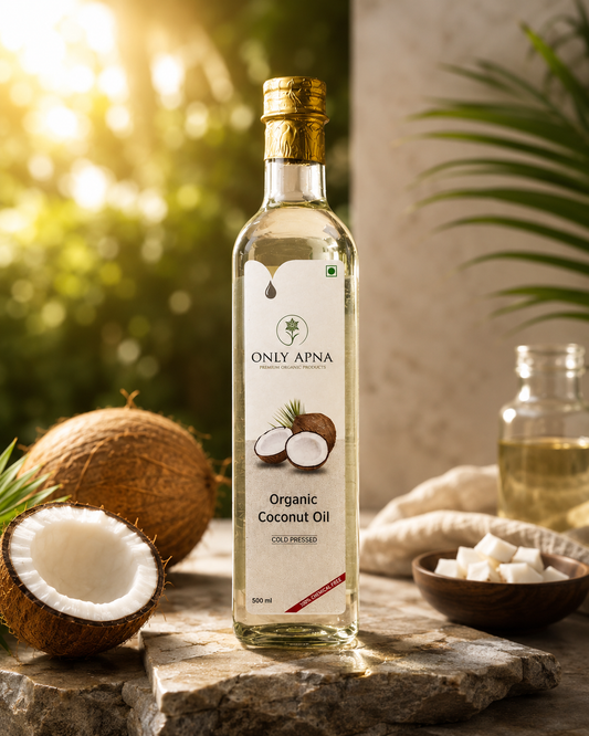 A bottle of ONLY APNA Organic Coconut Oil stands on a stone slab with whole and halved coconuts, a small bowl of cubed sugar, and a blurred tropical outdoor background with warm sunlight.